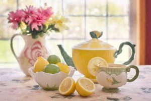 A close up photo of an afternoon tea setting in a house where we see the window behind. There's a floral tablecloth, a jug holding flowers, a yellow teapot, cup, and various lemons and limes. 