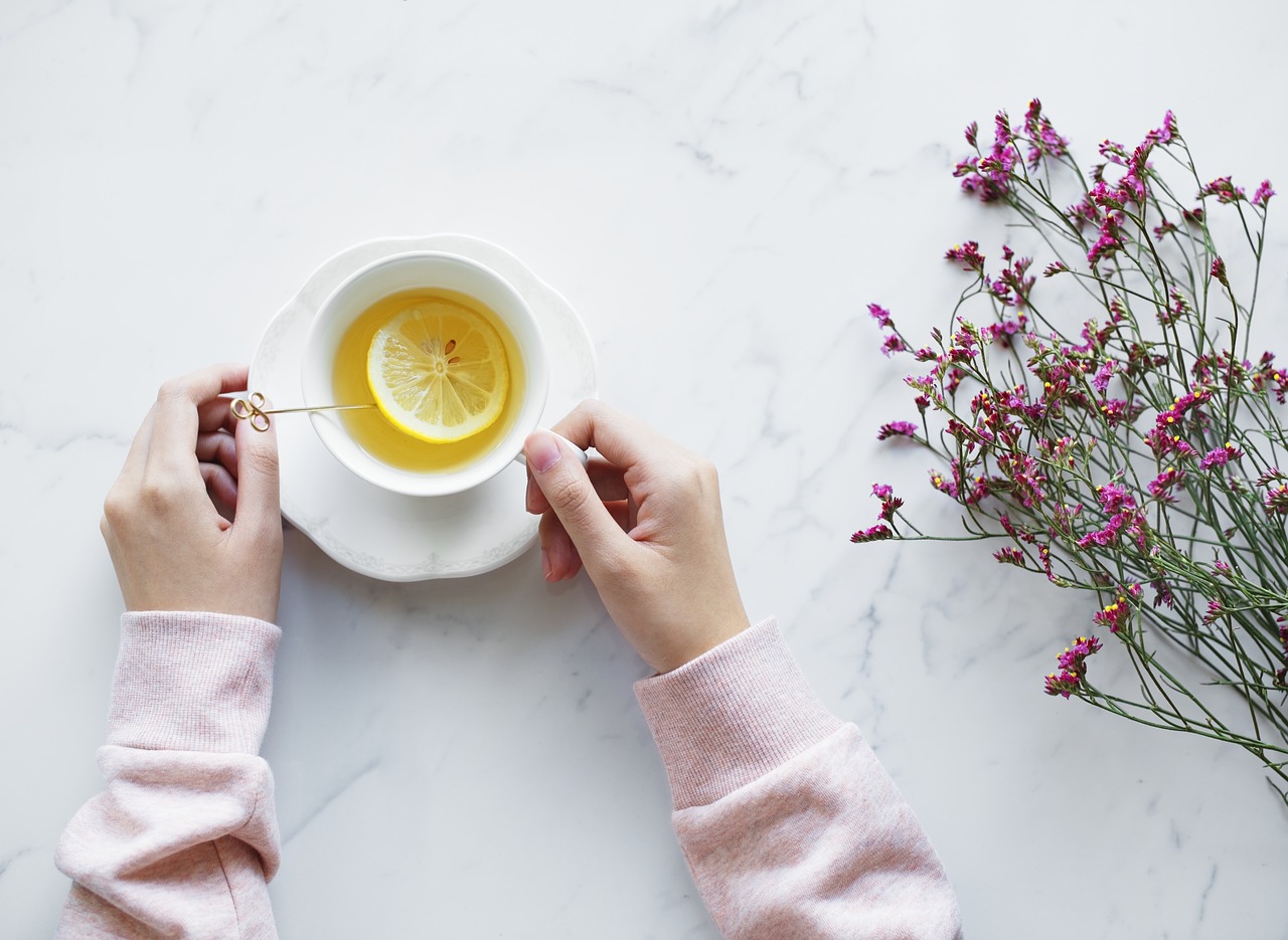 A birds eye view of a woman with her hands on her cup of hot lemon tea. It's sat on a white table and some purple flowers are lying to the right.
