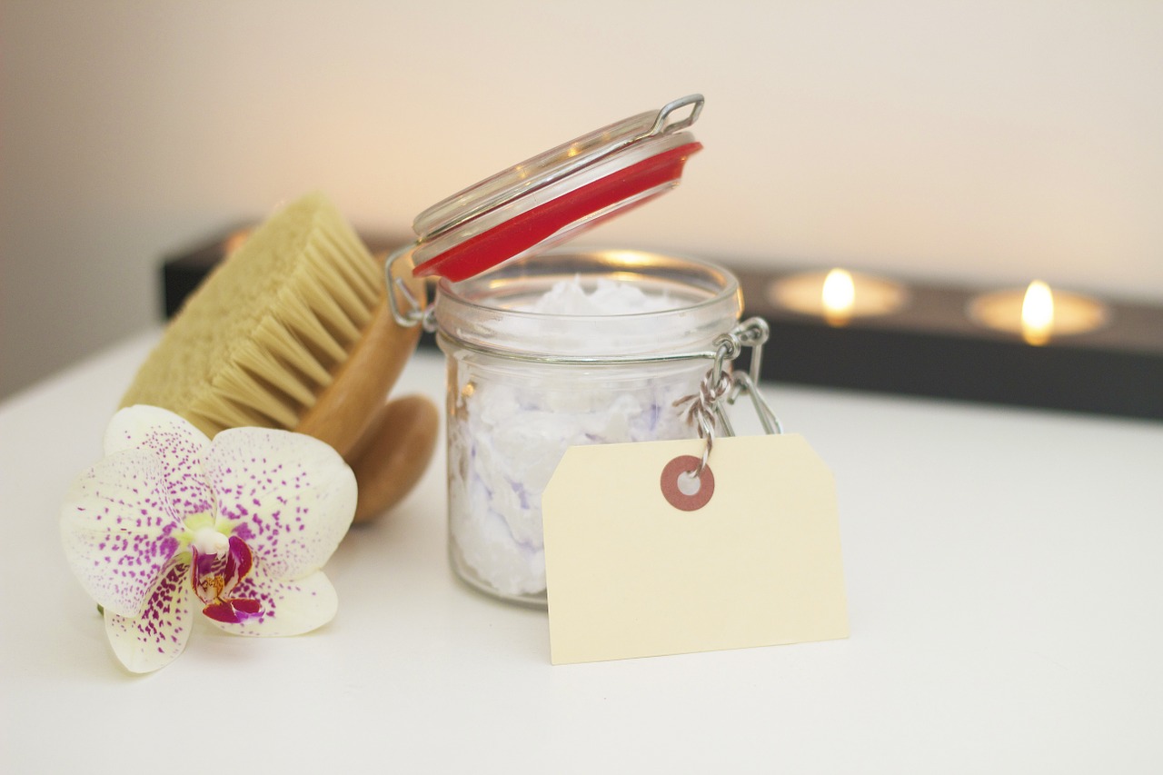 Bath salts in a small glass jar, along with an orchid flower and body brush sat on the white surface of a bath. Behind are tealights.