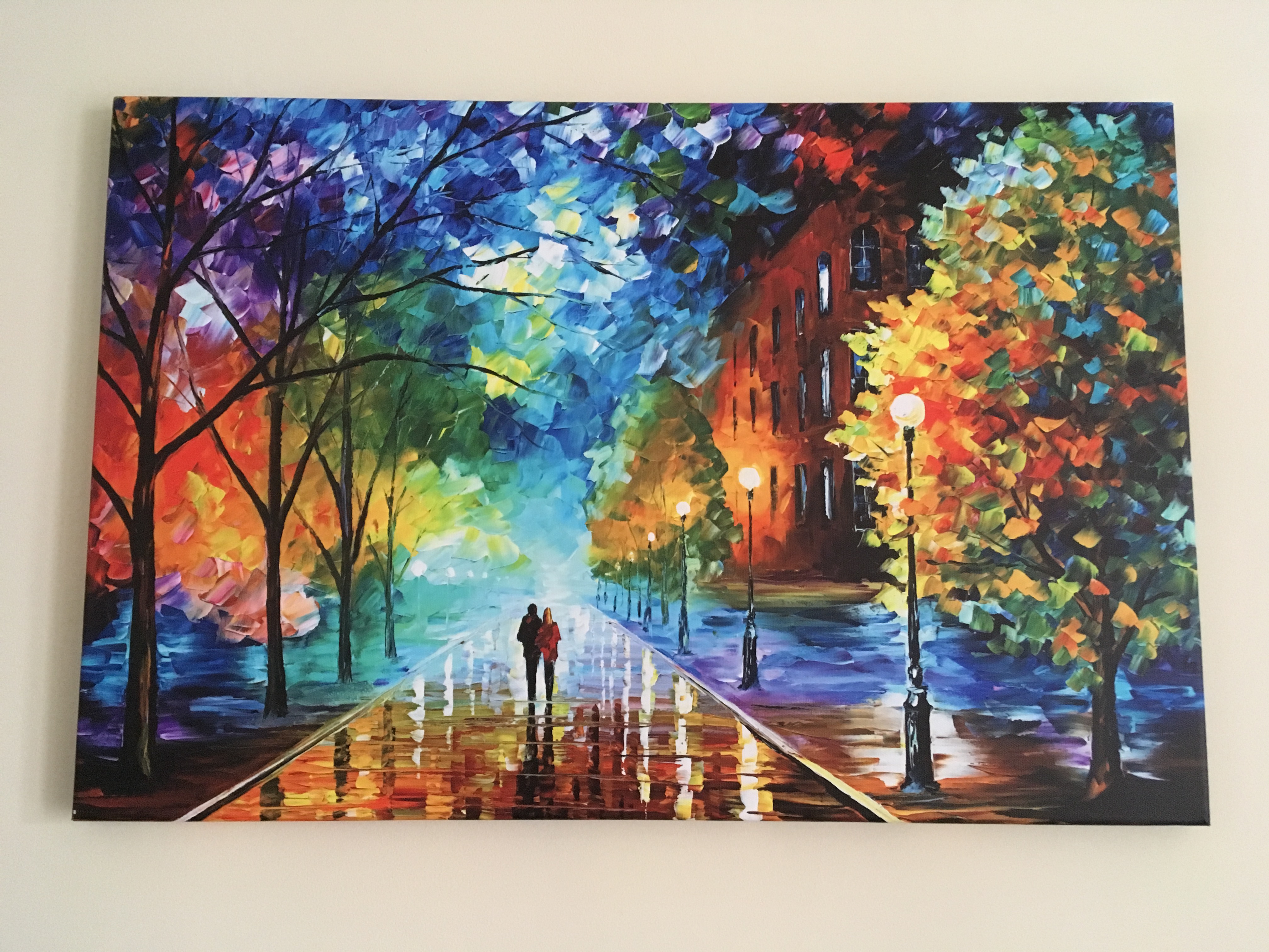 A photo of the art on the wall. It's a street scene with two people walking on the pavement in the distance, with trees to either side and buildings to the right. It's as though the ground is wet with rain as it glistens. The whole piece is done in a rainbow of colours and looks like an oil painting.