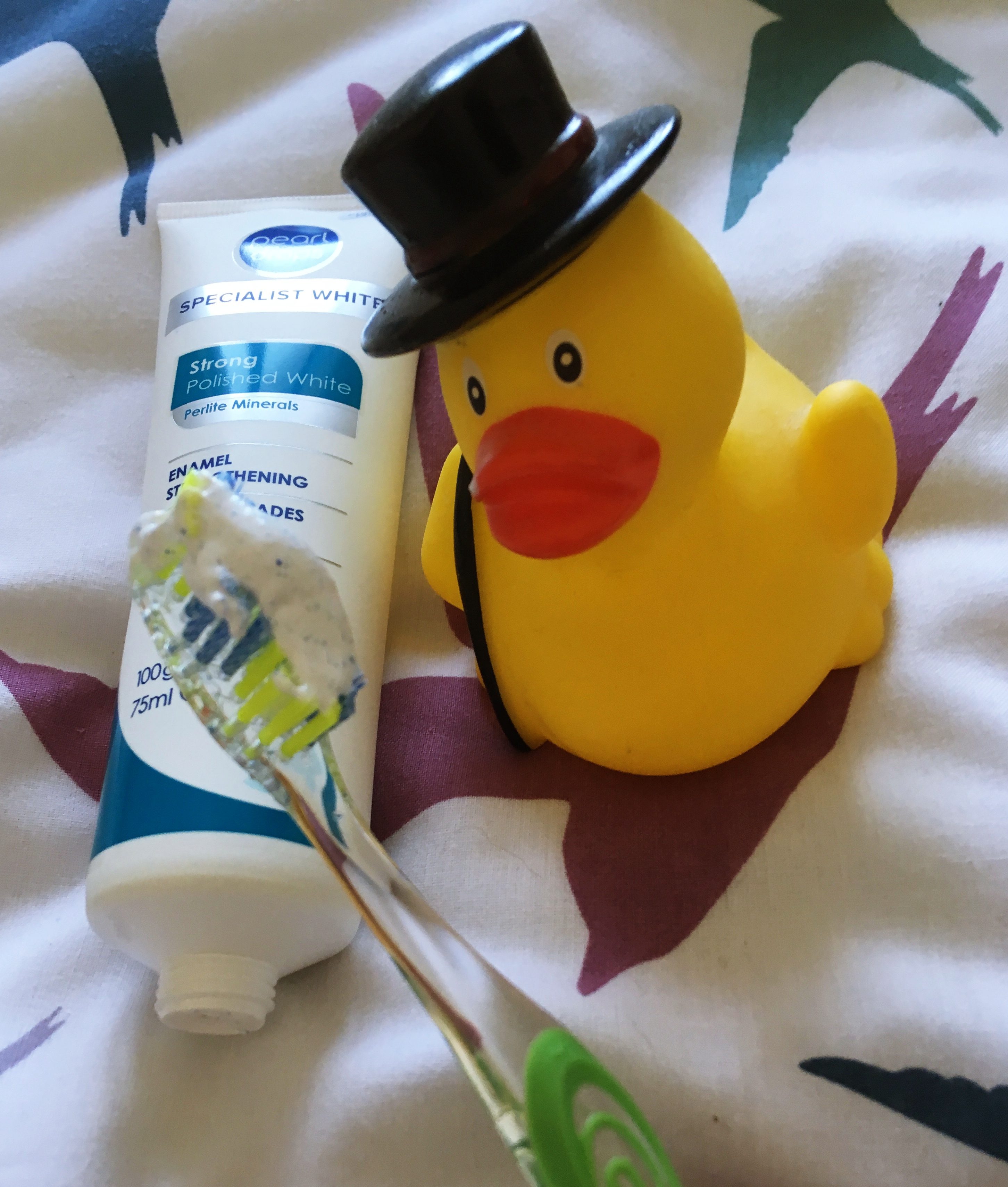 A photo of the toothpaste tube on my bed cover set next to a rubber duck, and my toothbrush to the front with a blob of the toothpaste on it.