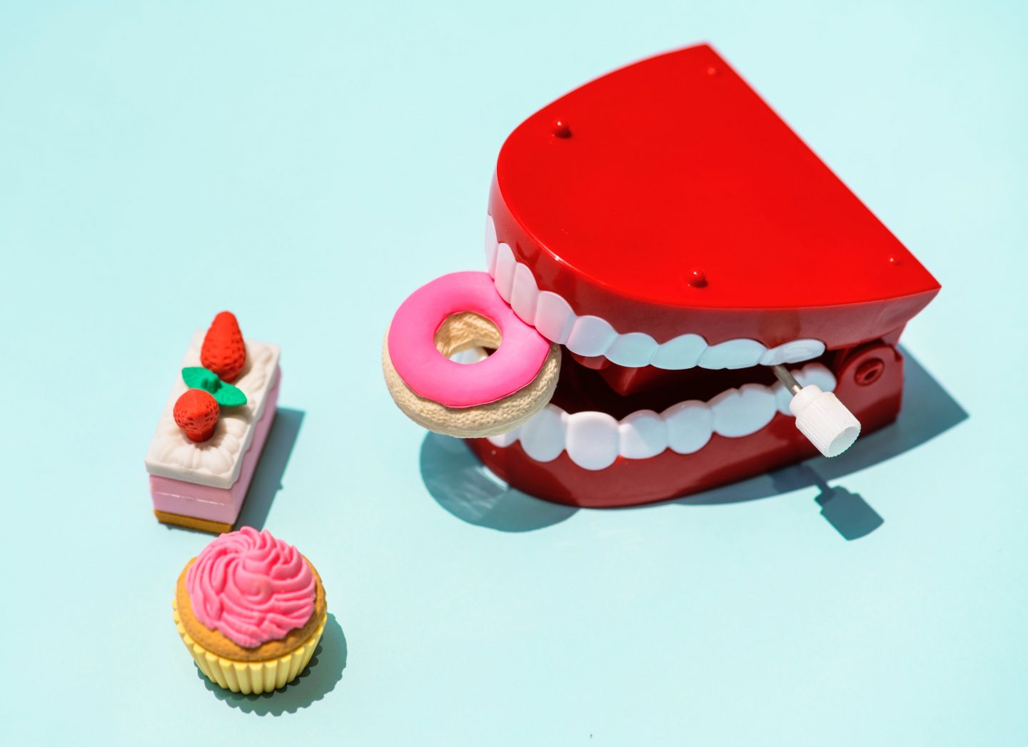 A wind up toy set of false teeth. They're holding a mini toy donut. There are two other tiny toy sweet treats next to the teeth. They're all set on a blue background.