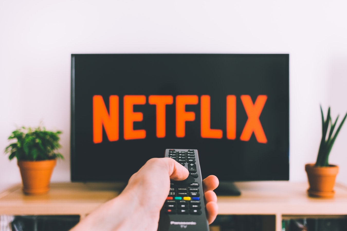 A hand reaching out with a remote pointed at a TV. On the TV is the Netflix logo.