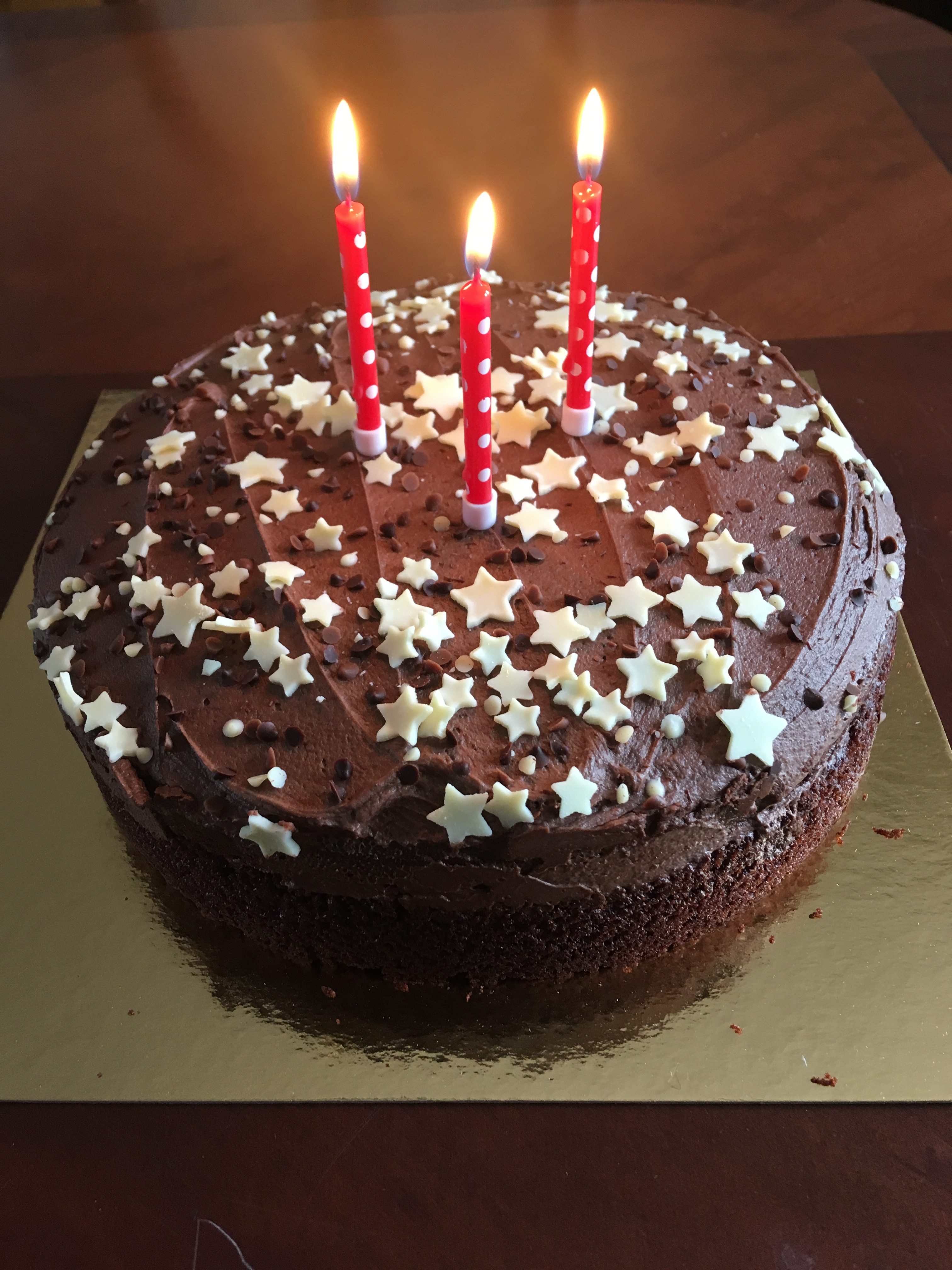 A photo of my birthday cake. It's a large circular chocolate cake, with white star chocolate sprinkles over it. There are three red lit candles.