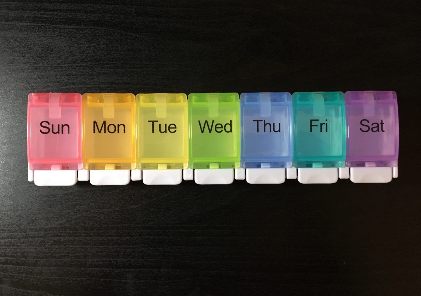 The Auvon pill box on my black desk. It has 7 compartments with a day of the week written on each. It's a long rectangle in white, and the lids for each day are in a different colour.