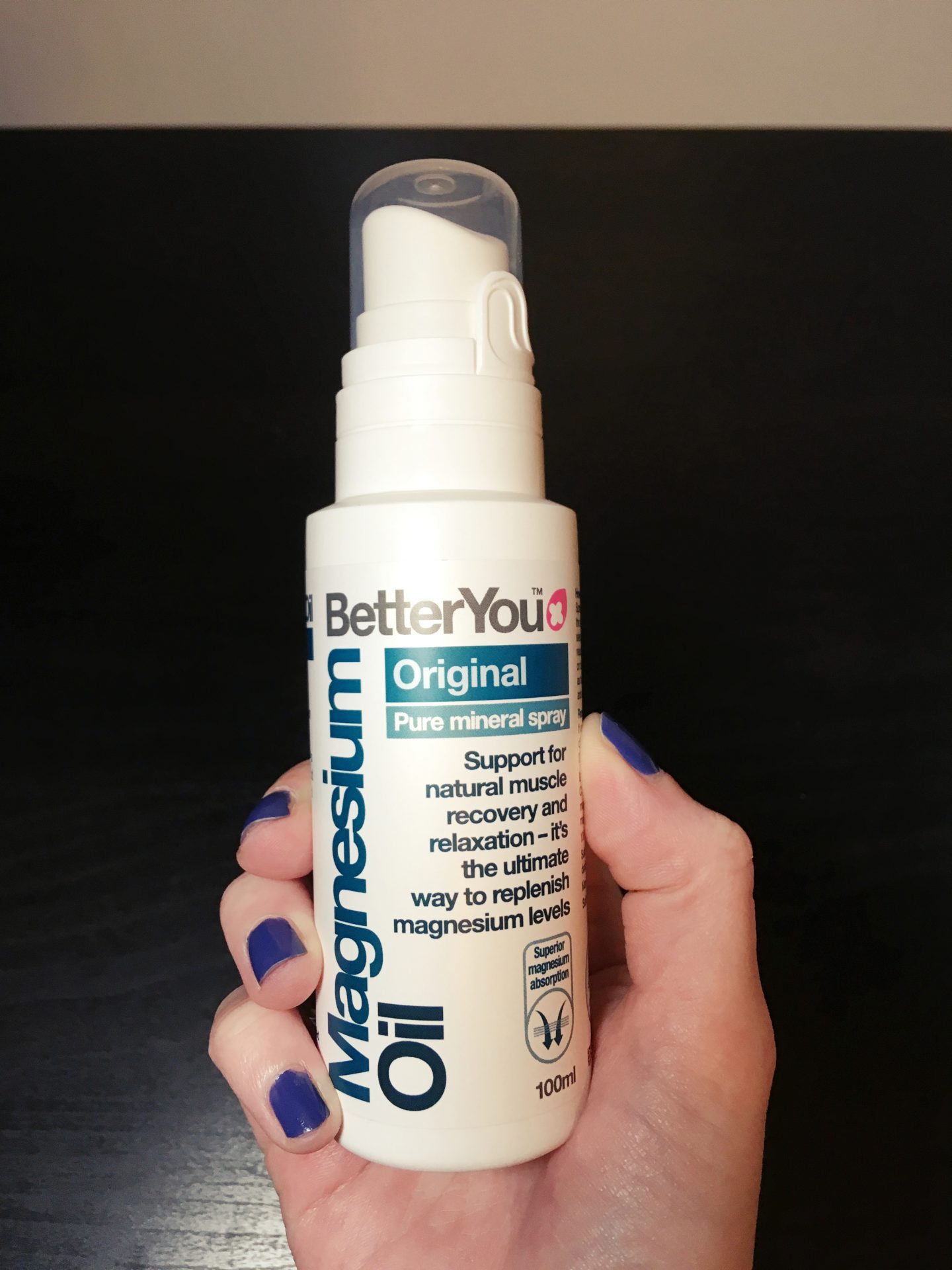 My hand holding the BetterYou Magnesium Oil Spray against a Black background.