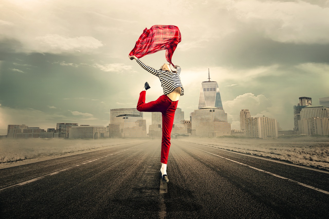 A woman standing on a road with a city scape behind her. She's stretching and holding a top above her head, as though she's free and feeling liberated.