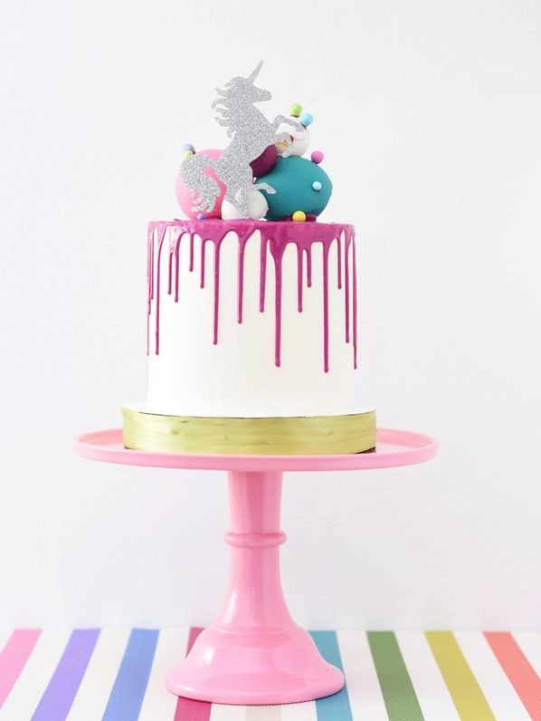 Photo of a cake on a stand, which is on a rainbow striped tabled. The cake is large with pink icing, and a silver unicorn on top.