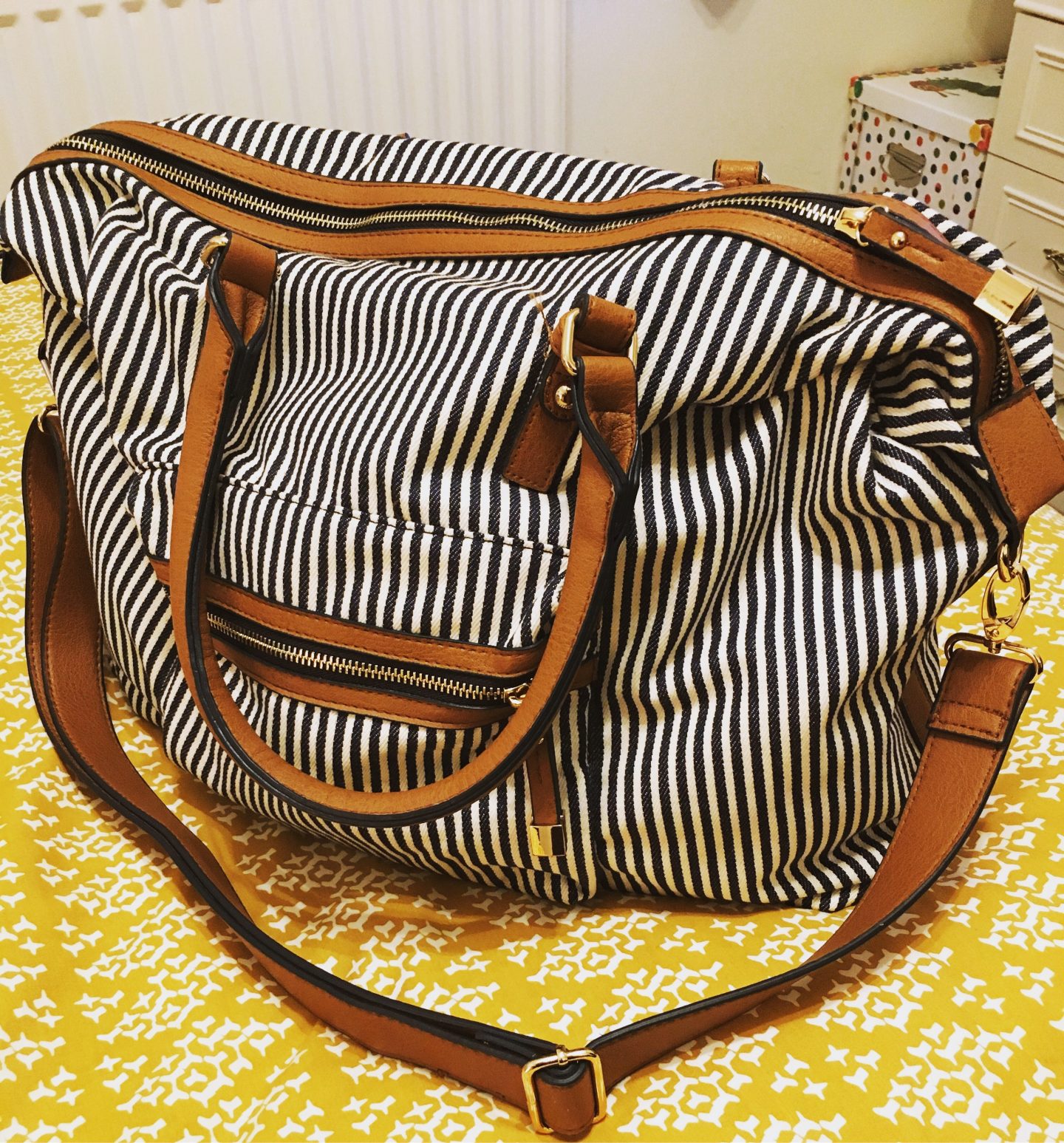 A photo of my new weekend travel bag, which is black and white striped with brown detailing and brown straps. It's sat on my yellow duvet in my room.