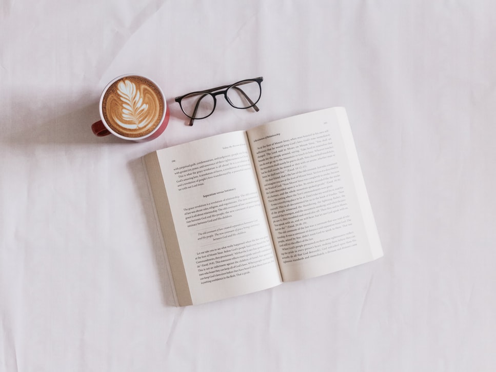 A photo of a white background with an open book, pair of glasses and cup of coffee. The photo is taken as a birds eye view.