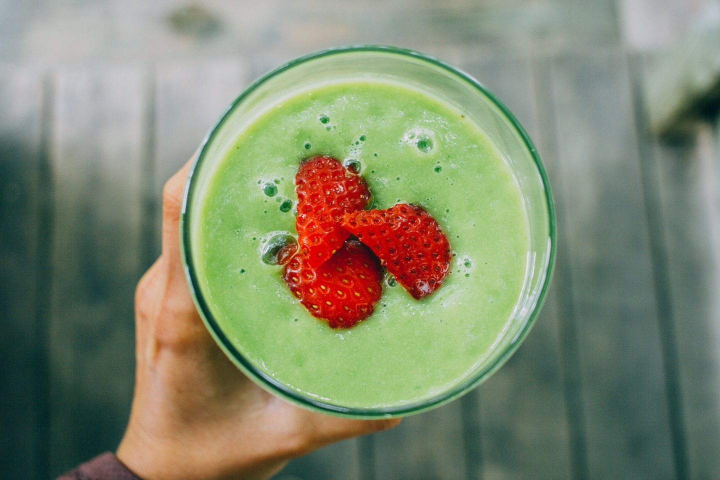A birds-eye view of someone holding a glass. We see the green, thick smoothie and some chopped red strawberries in the middle of the drink.