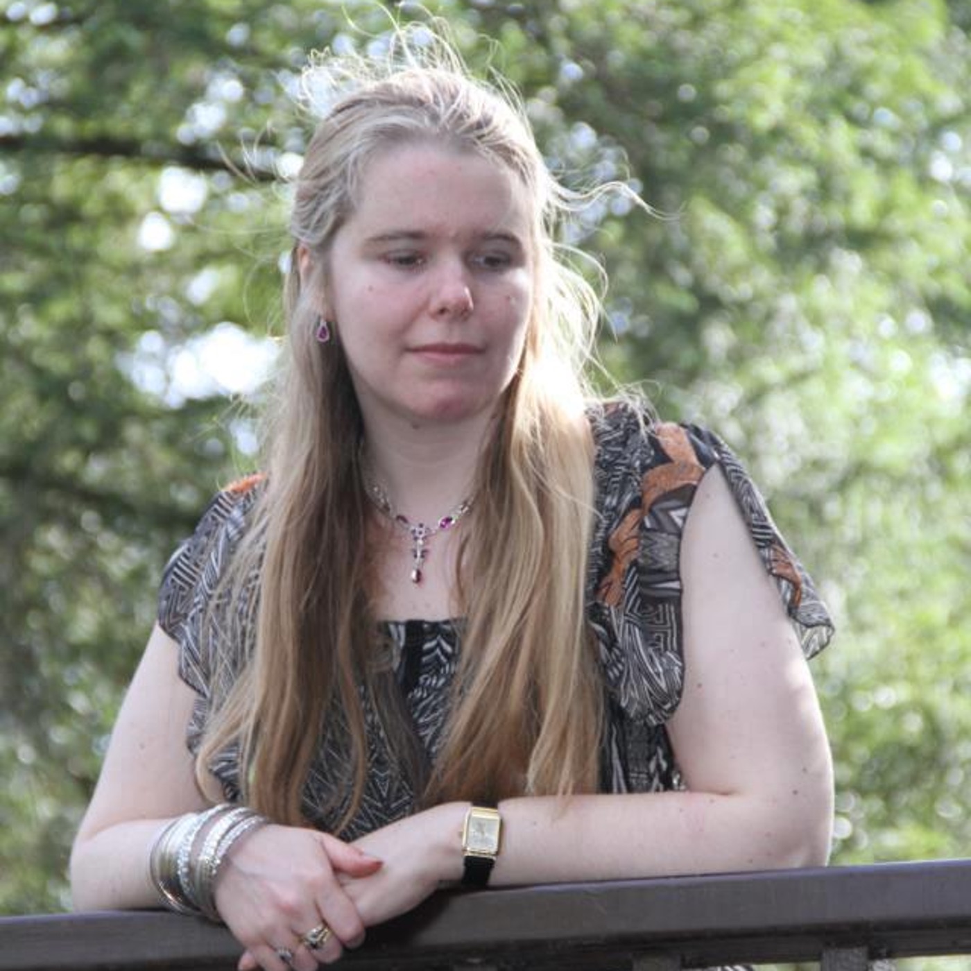 A photo of Kirsty where we see her upper body as she leans on the rail of a bridge. She has long blonde hair, she's wearing a short sleeved top, and she has pretty silver bracelets and a necklace.