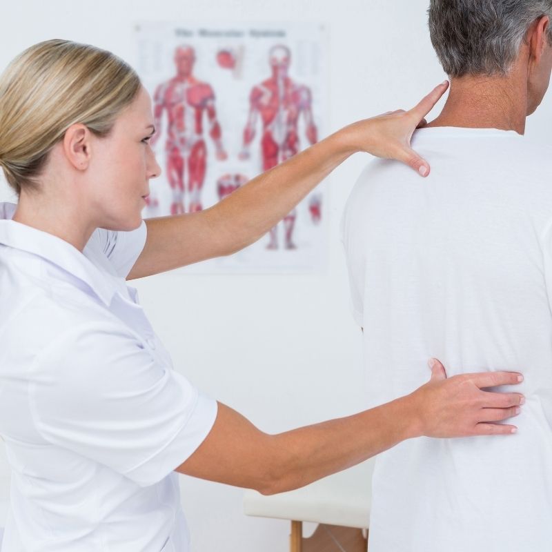 An osteopath in white is putting pressure with both hands on a man's back to help treat chronic pain as he stands upright. On the wall behind them is a poster of the human body.