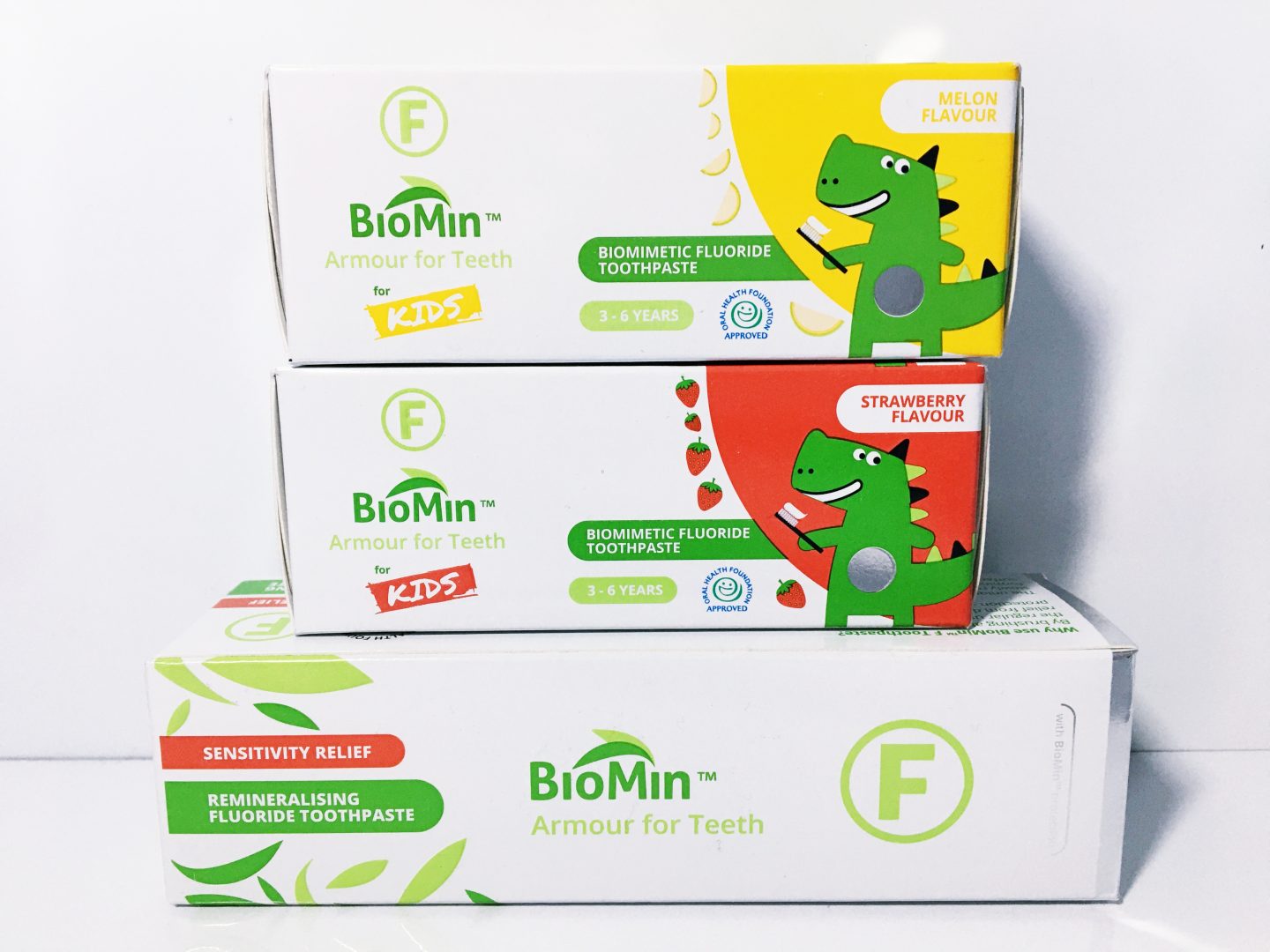 A photo of three BioMin toothpaste boxes, which are white, green, yellow and red in design, on a white desk against a white background. The boxes include the BioMin F toothpaste and the two toothpastes for kids, all gifted for this review.