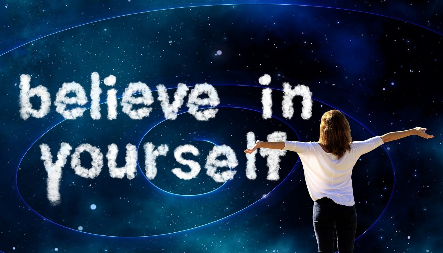 A dark background like a night sky with stars. A woman stands to the right in a white t-shirt with her arms stretched out to suggest confidence and her back to the camera. On the sky are the words "believe in yourself" made to look like clouds.
