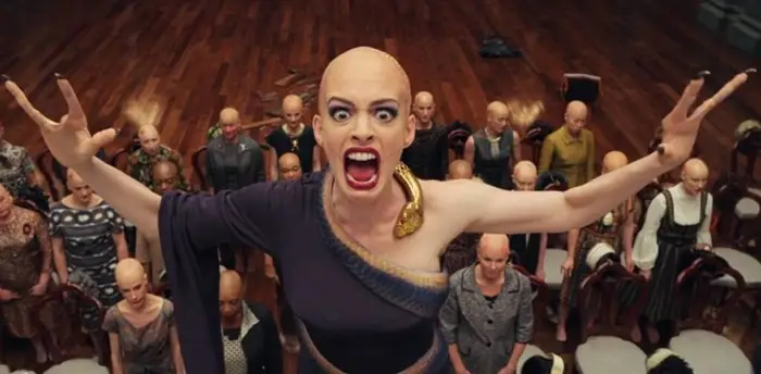 A still from The Witches movie. In the background are many bald people sitting and standing. Anne Hathaway is standing in front looking directly forward. She's wearing a one-sleeved outfit, her arms raised in the air as she screams with her mouth open and teeth bared. Her hands are deformed, with only two long fingers spaced apart. She'd bald with scars on her head.