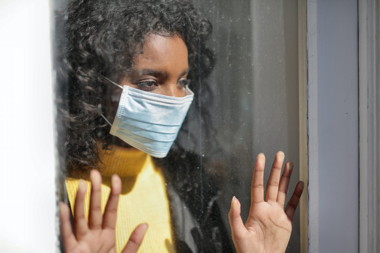 A woman in a yellow top, with dark curly hair and dark skin is looking out the window; she's wearing a mask and has her hands up to the glass as though in lockdown during the pandemic.