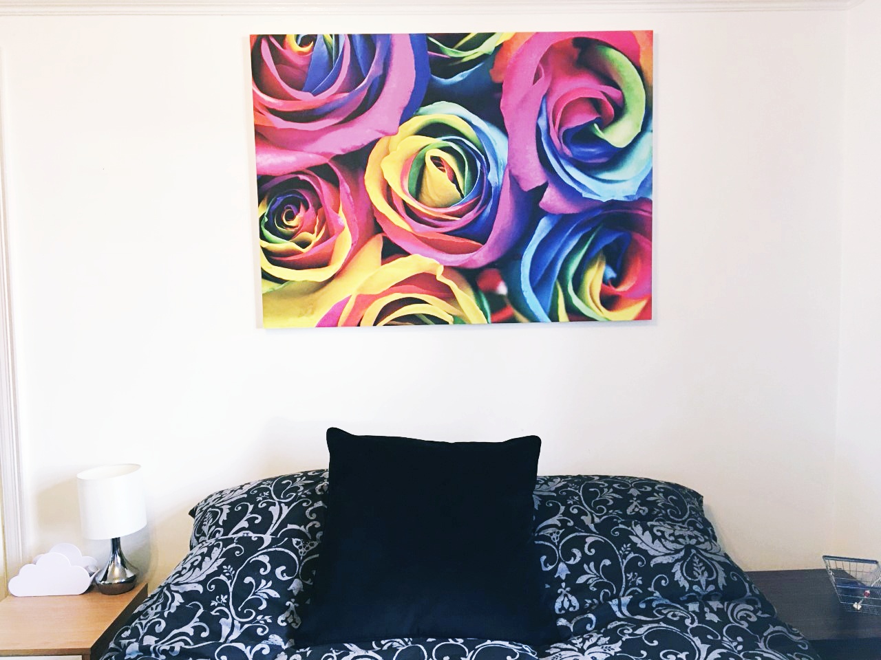 A photo taken in my room of the wall where the Photowall canvas has been hung. There's a double bed with the pillows against the wall, with a black and grey damask duvet cover. There are bedside tables to either side. The canvas is large, though not quite the width of the bed. It features colourful rainbow roses.