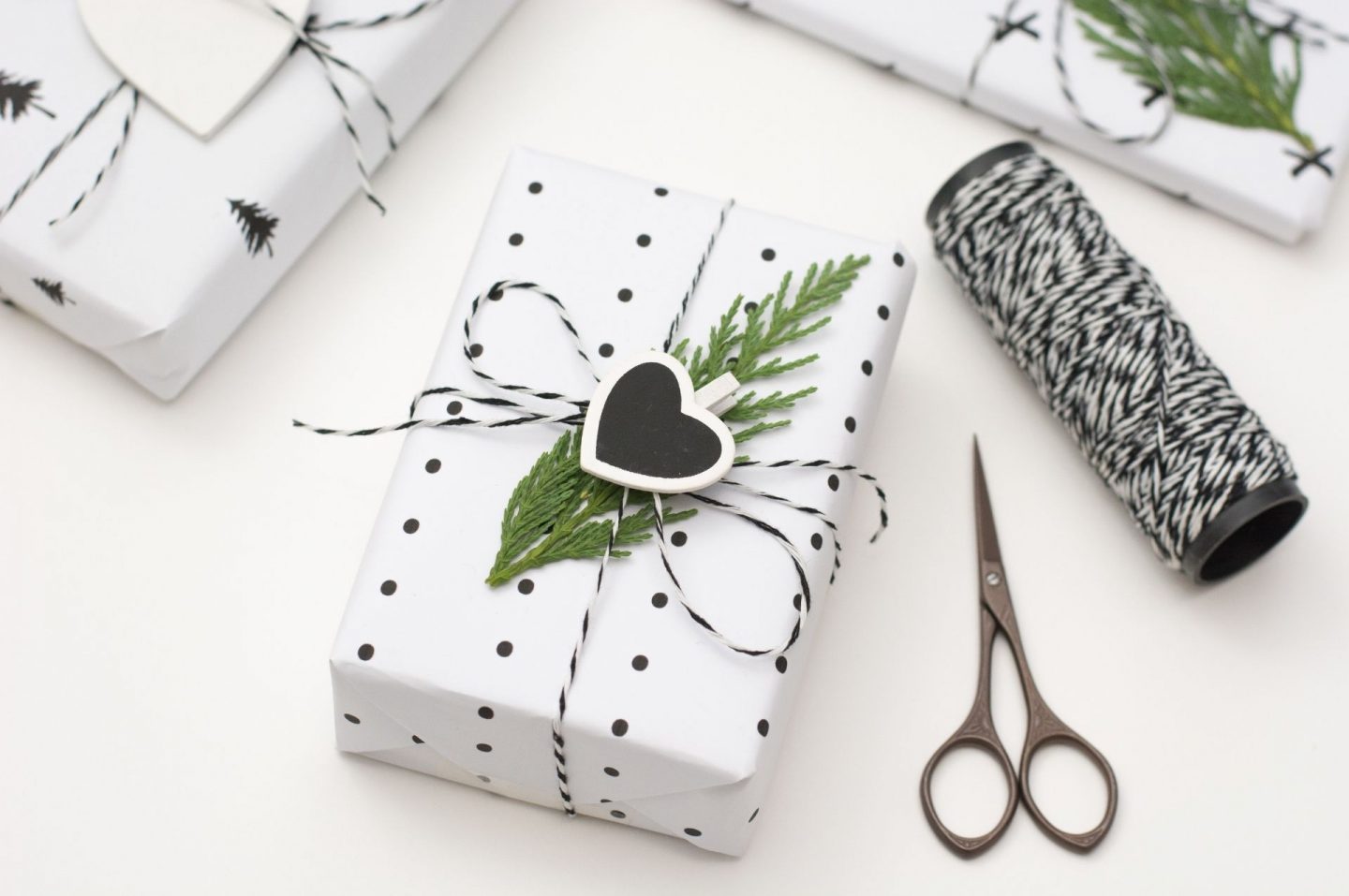 A photo of Christmas gifts that are being wrapped in black with black polka dots and Christmas tree paper. There's a black and white ball of twins and a pair of scissors next to the gifts.