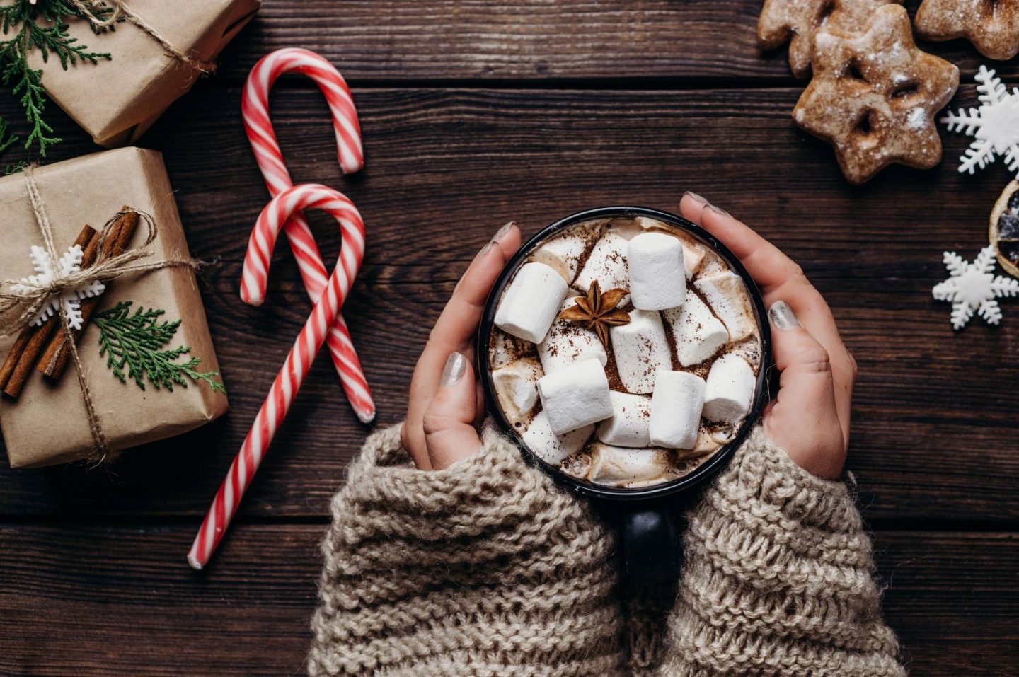 A birds-eye photo of a wooden desk with Christmas things on it, like two candy canes, sugar cookies and wrapped gifts. A woman has her hands wrapped around a mug of hot chocolate with marshmallows.