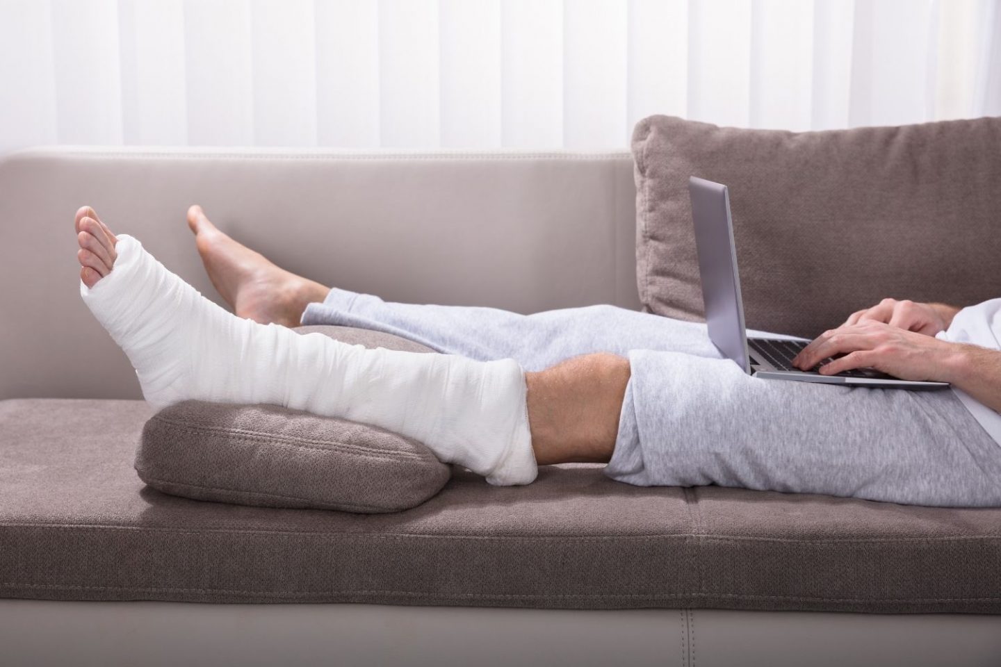 A photo of a man's legs as he lies on a sofa with a laptop on his thighs. One leg is propped up in a bandage as though he's broken his ankle. This is for the health news story of a new biomaterial for bone breaks.
