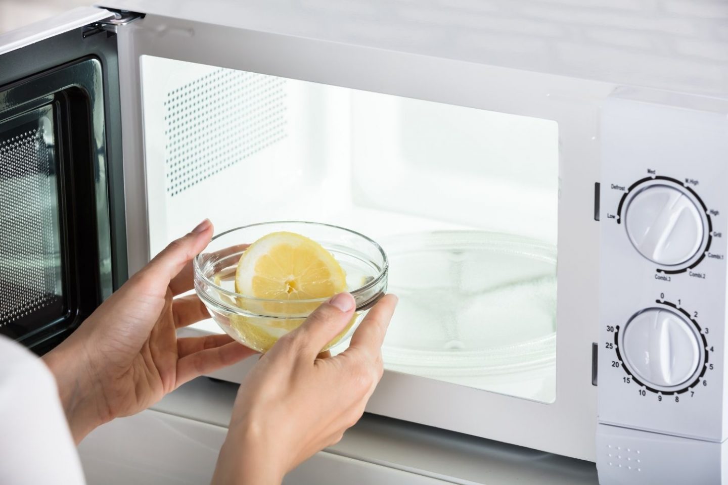 A close-up photo showing a white microwave with the door open. A woman is placing inside a clear bowl with water and half a lemon. This is the cleaning hack for getting rid of odours and cleaning a microwave.