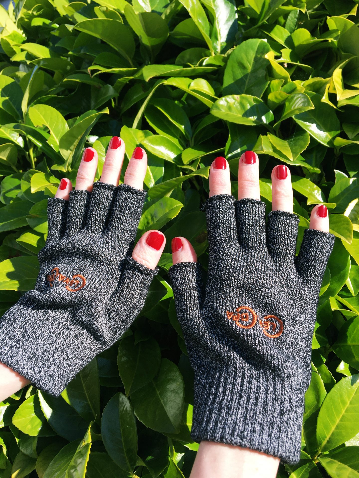 A close-up photo of my hands wearing the grey fingerless knit gloves from Copper Clothing that I was kindly gifted for this review. My hands are held up with the green leaves of the plant in my garden as the background.