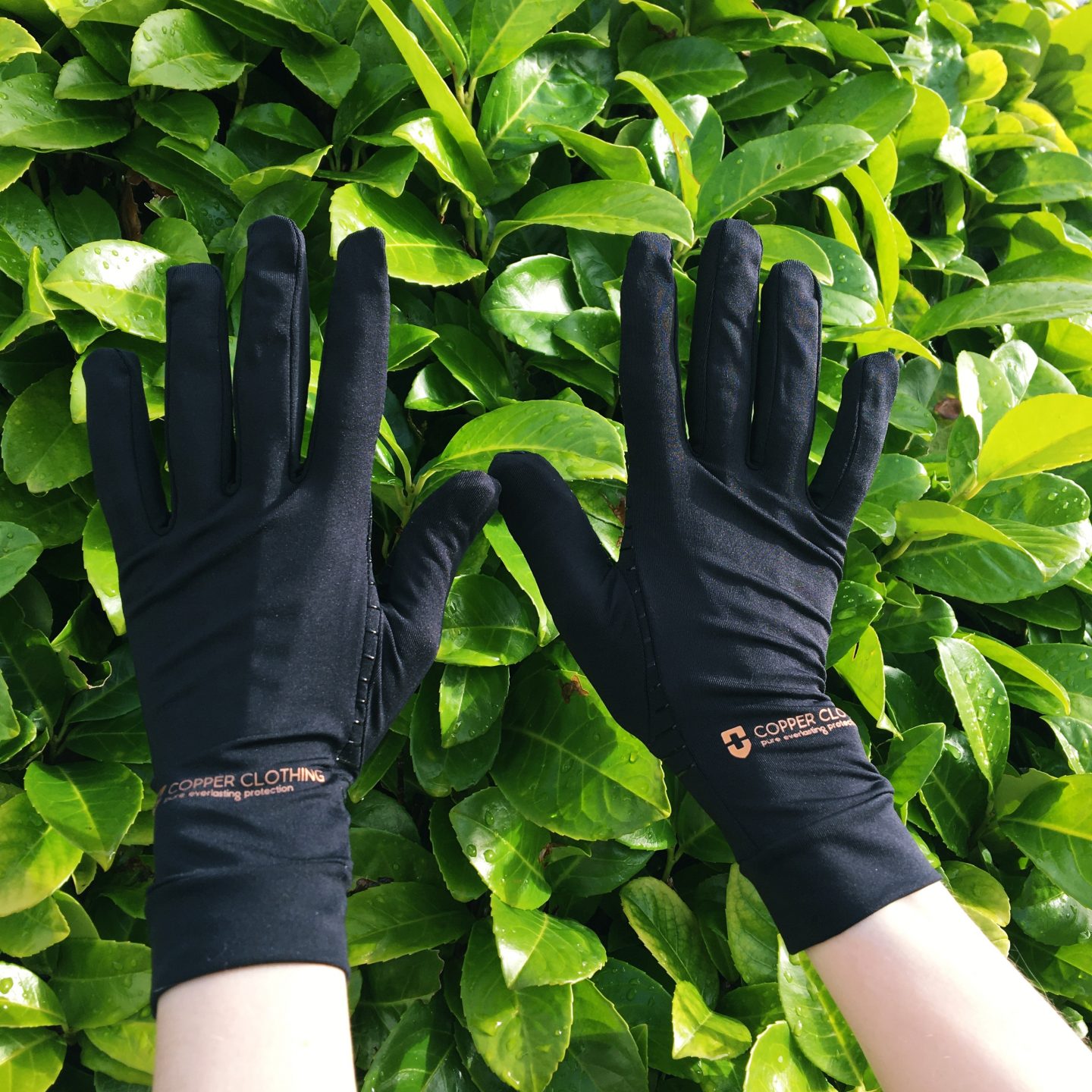 A close-up photo of my hands and forearms. I'm wearing the black Copper Clothing compression gloves I was gifted for this review. My hands are held up with the green leaves of the plant in my garden as the background.
