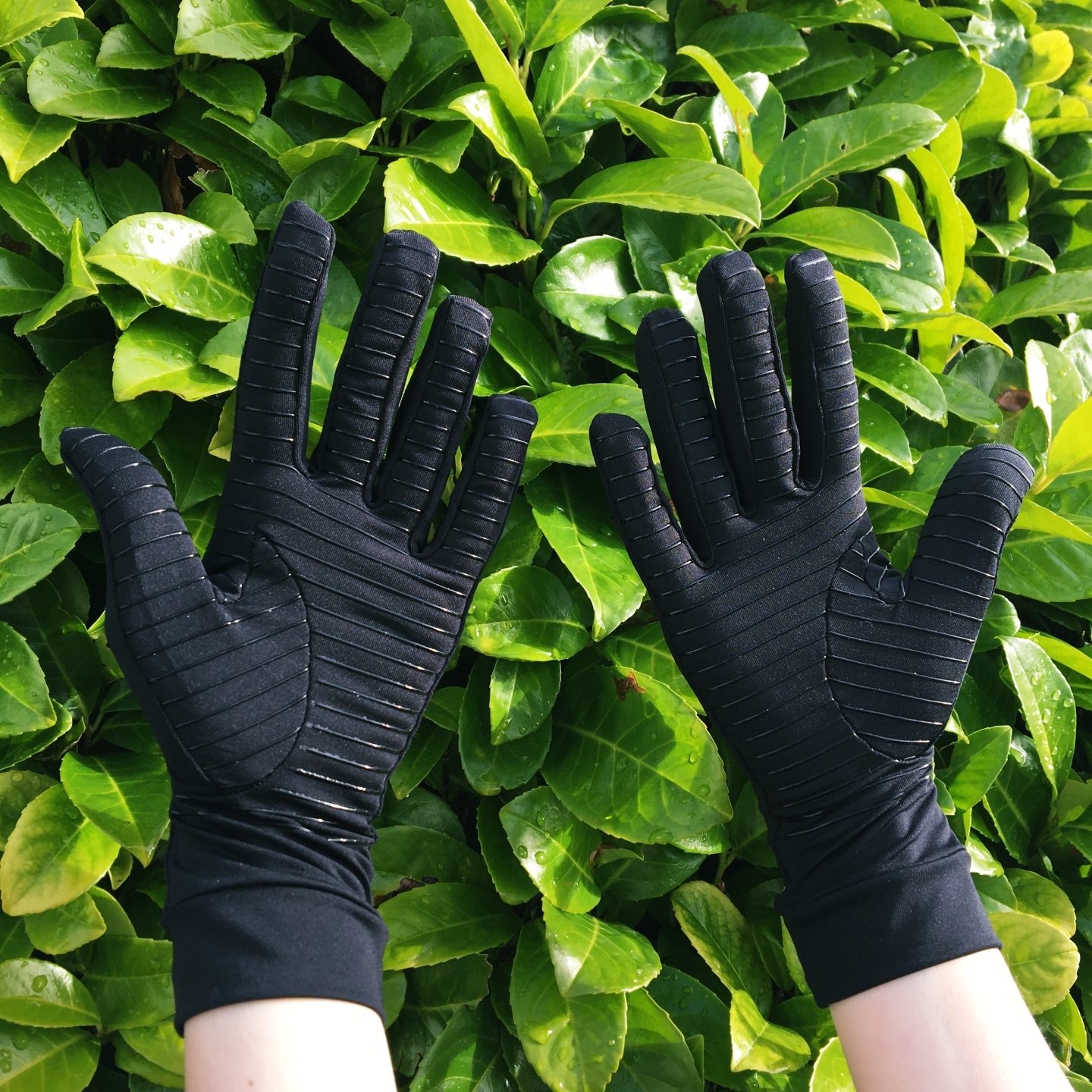 A close-up photo of my hands and forearms. I'm wearing the black Copper Clothing compression gloves I was gifted for this review. My hands are held up with the green leaves of the plant in my garden as the background. My hands are turned to show the palms, where lines cross over the palms and fingers to provide a rubberised grip.