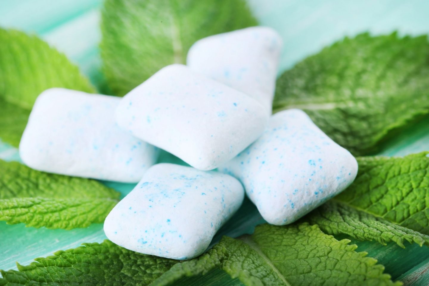 A close-up photo of five pieces of minty sugar free chewing gum sat on top of mint leaves.