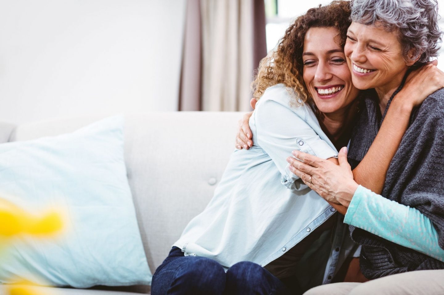 A photo of two women on a grey sofa hugging and smiling. One looks to be in her 30s or early 40s, and the other is perhaps her 70s, so they could be a mother and daughter or a woman with her carer.