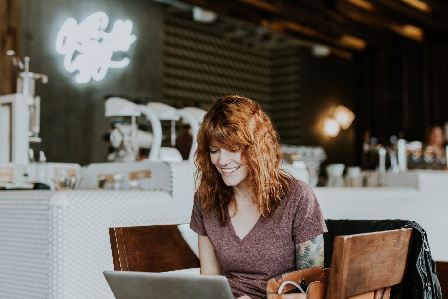 A photo of a woman, perhaps in a coffee shop, smiling and looking down at her laptop.