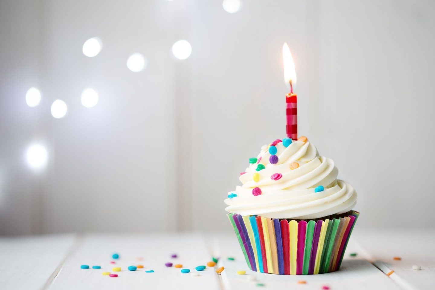 A photo of a cupcake in a rainbow coloured case with colourful sprinkles and a single red candle. It sits on a white table with a white background, with a few dots of light behind it.