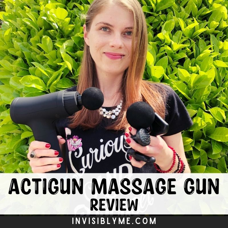 A photo of me in the garden against a green tree holding the Actigun massage gun for this review in one hand and the other head attachments in the other. I'm wearing an Alice In Wonderland black t-shirt. Below is the title : Actigun Massage Gun Review.