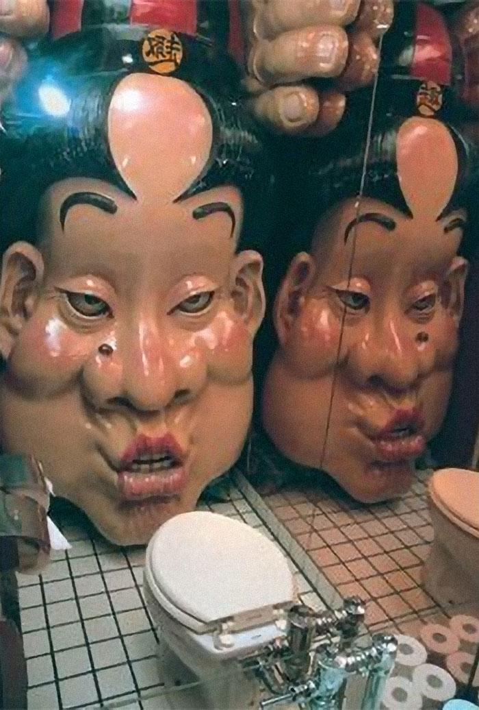 The bathroom in Japan, with the photo from Bored Panda for this funnies post. It's a small stall with a toilet and mirror to the right. In front of the toilet is a giant plastic face of a man pouting his lips. It's as tall as the room itself.
