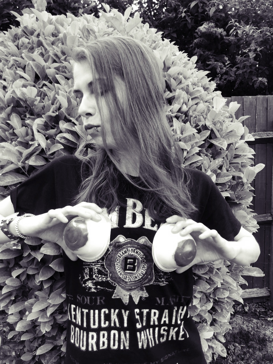 A photo of me in the garden in black and white, wearing a t-shirt and holding up two yoghurts (white with a red part at the bottom which looks like a boob). I've got my head tilted to the side and I'm pouting.