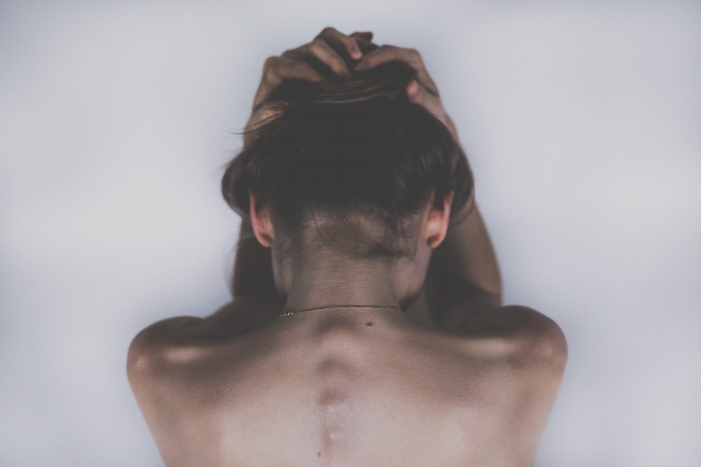 A darkly coloured image showing the upper bare back and shoulders of a woman. Her hands are on her head and she faces away from the camera, suggesting she's suffering, perhaps with mental health, illness or chronic pain.