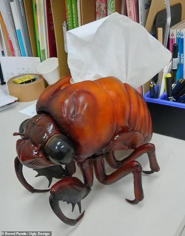A giant shiny brown bug with six legs and pincers at the front with big bulging eyes. Along its back is an opening with a tissue popping out. It's sat on a desk by books, showing it's a large size.