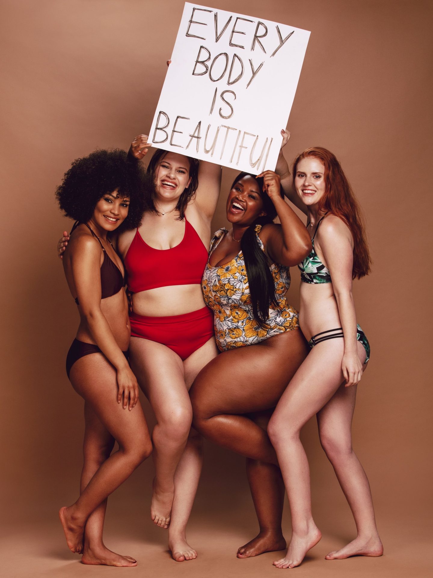 A dark beige to brown background. Four women are stood up wearing swimwear. They're holding a sign above their heads reading "Every body is beautiful". They're all smiling and they're all different, like different sizes, hair styles, skin colour, body shape and so on.