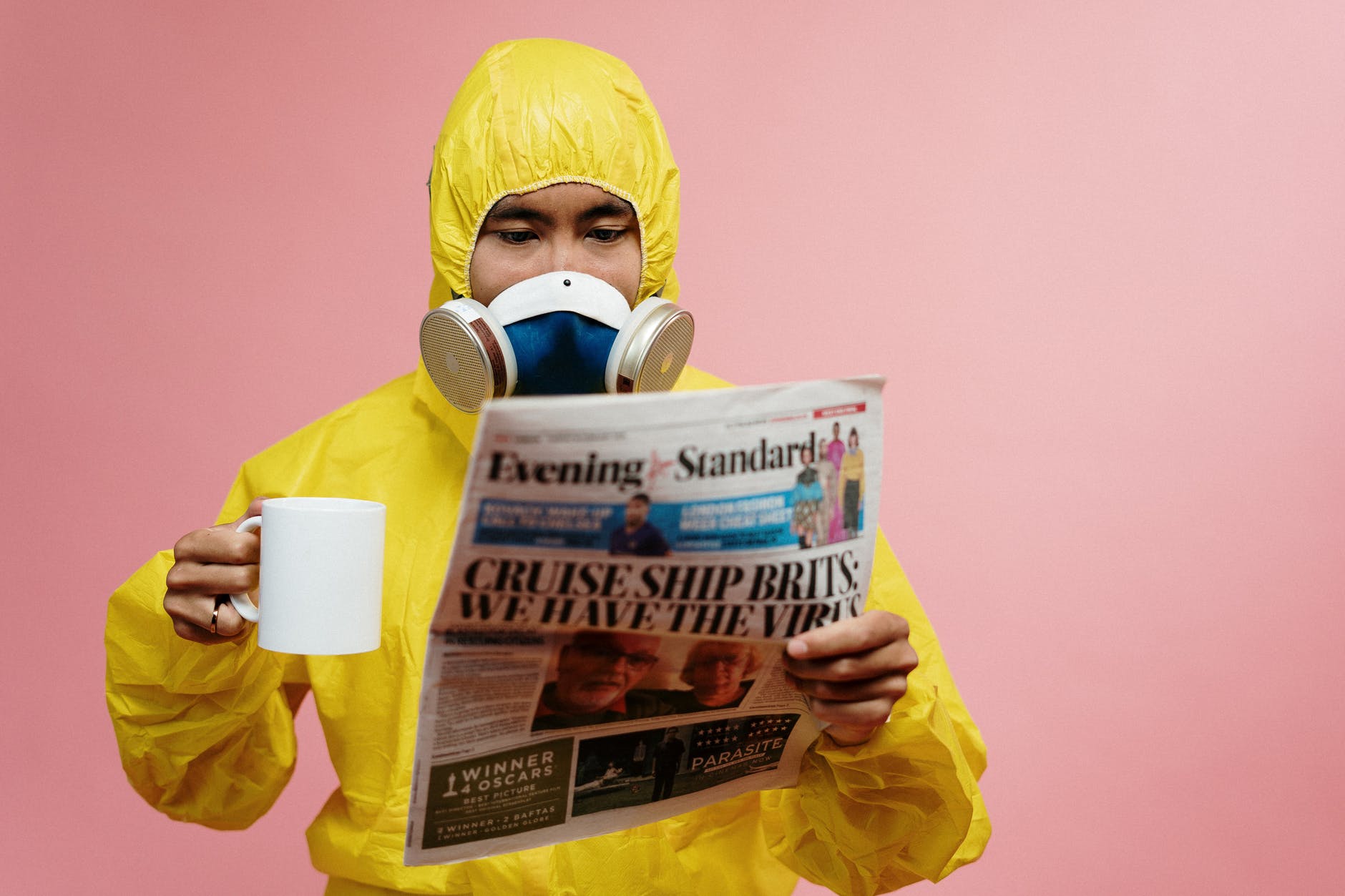 A person holding a mug and a newspaper, dressed in a yellow hazmat suit to suggest reading the news during Covid19.