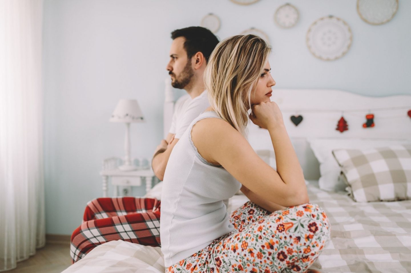 A man and woman sat on their bed facing opposite directions. They both look fed up as though they've had an argument or their personal boundaries have been disrespected.