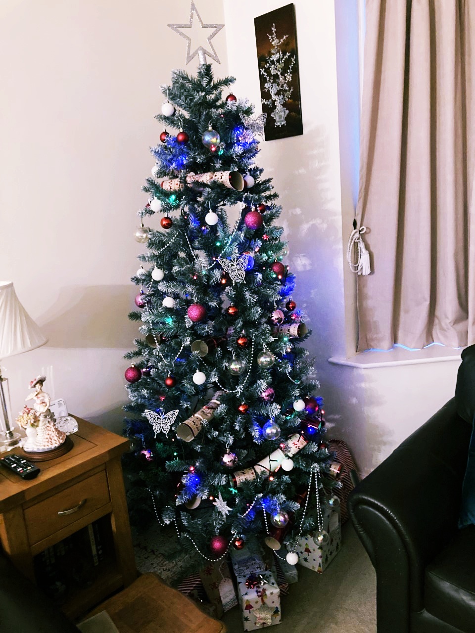 A photo of our Christmas tree, decorated with baubles, sparkly trees and butterflies, and crackers. It's in a corner of the living room.