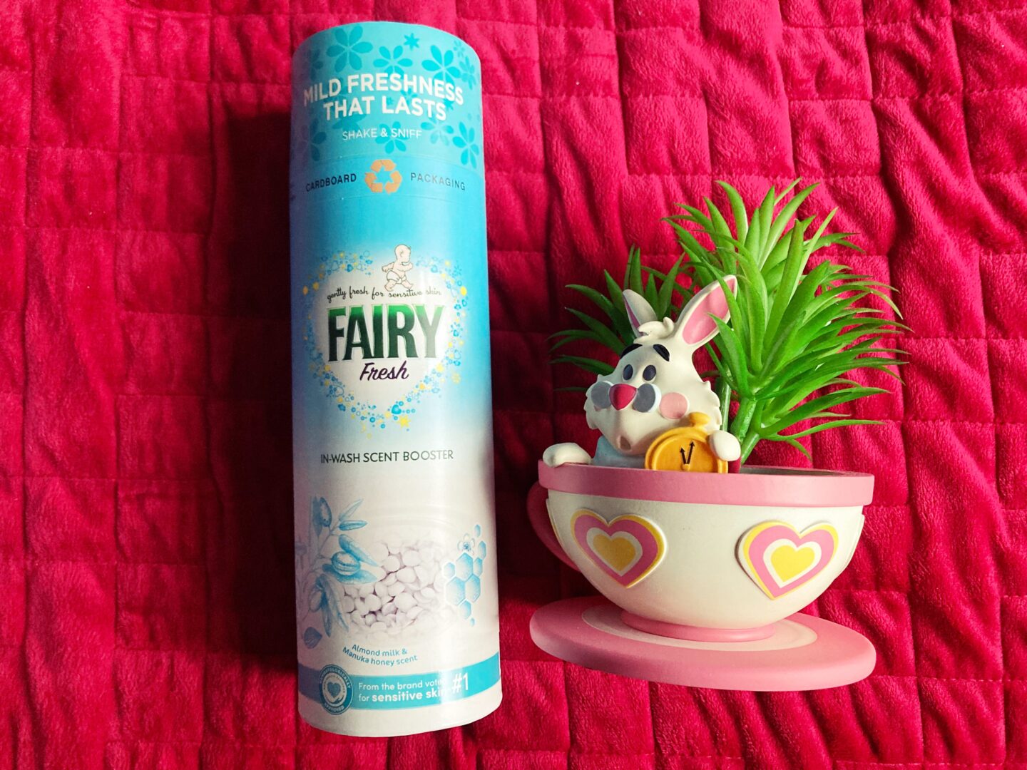 A photo of one of the Fairy Outdoorables products against a red blanket. A teacup and saucer-style potted plant in the style of Alice In Wonderland with the White Rabbit in it sits to the right of the product.