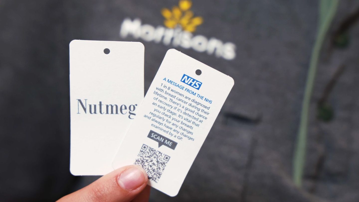 Two white labels are shown being held, one with the Nutmeg branding on and the other with the NHS message saying "1 in 8 women are diagnosed with breast cancer during their lifetime". There's a QR code underneath.