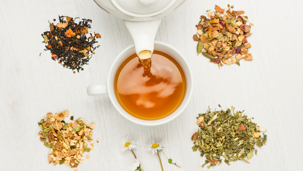 bird's eye photo of a cup of tea being poured from a teapot, with little piles of different tea leaves around the cup on the table.