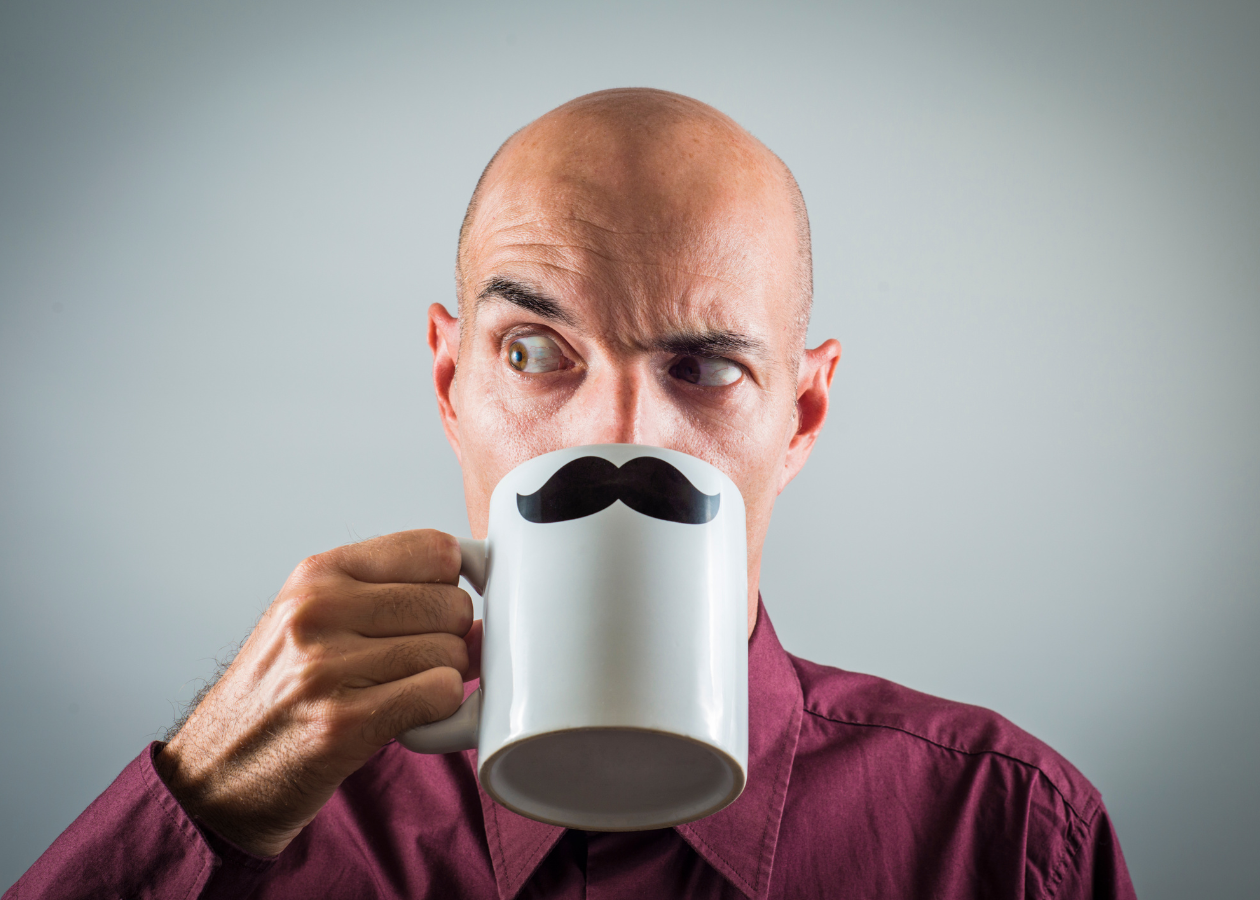 A man against a grey background is holding up a mug of tea with a moustache on the front of it. It's held up to his face to look like the moustache is on him and he's got an eyebrow up in a quirky appearance.