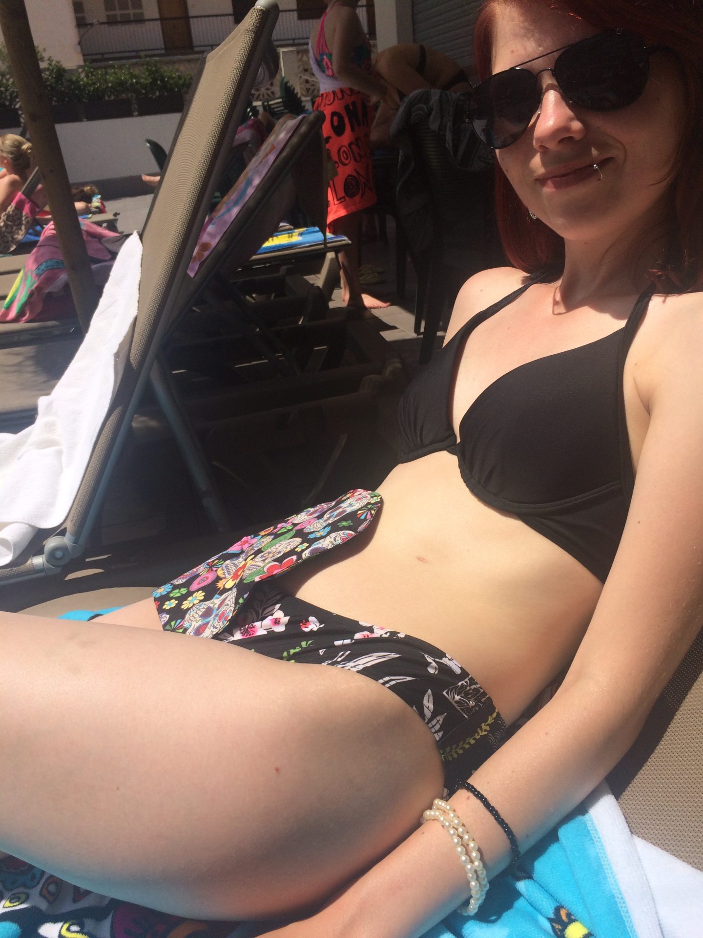 A photo of me reclining on a sun lounger wearing a black bikini, sunglasses and a sugar skull patterned stoma bag cover.