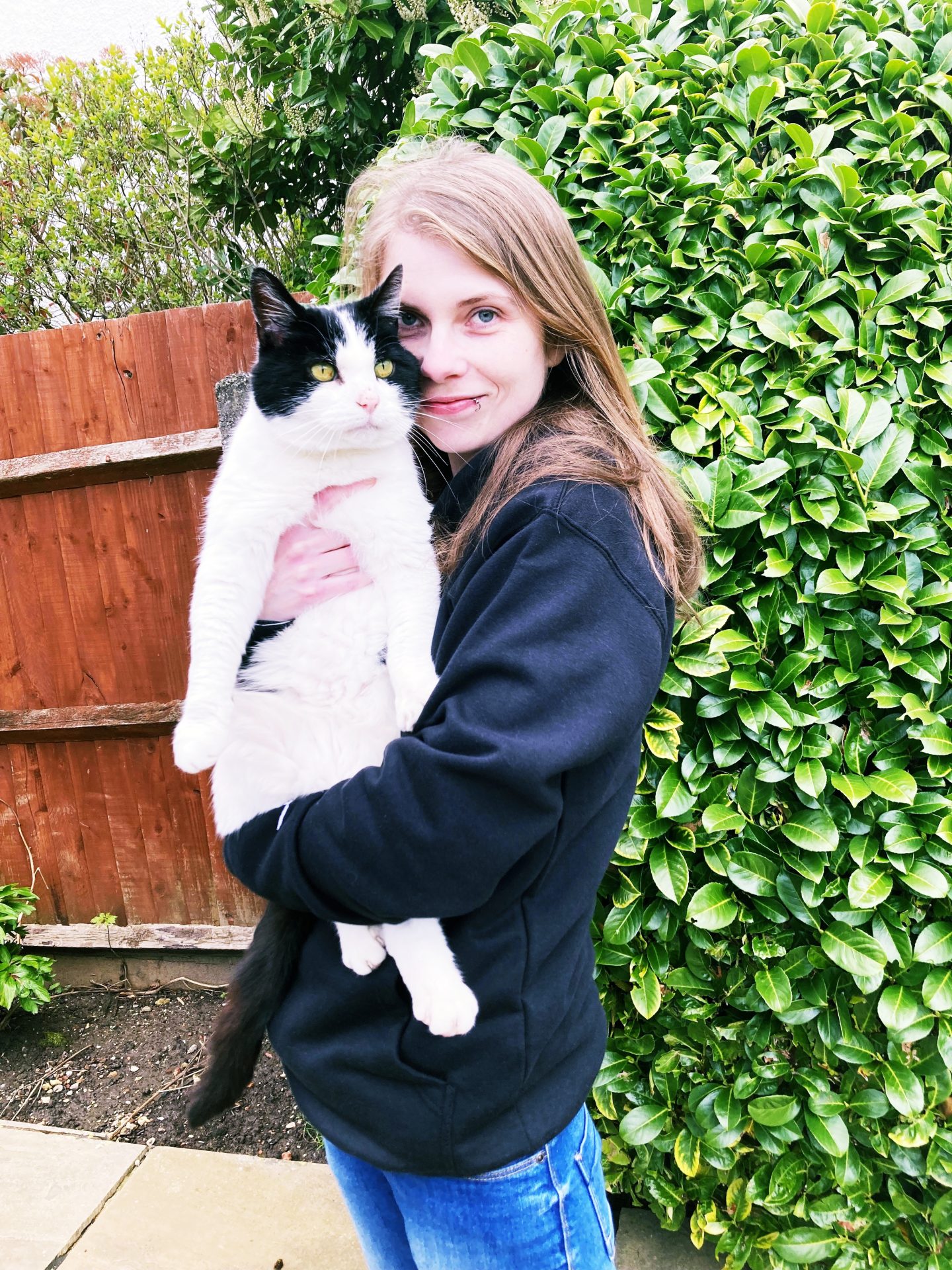 A photo of me in the garden wearing jeans and a black hoodie, holding up and hugging my black and white cat Virgil to my face.