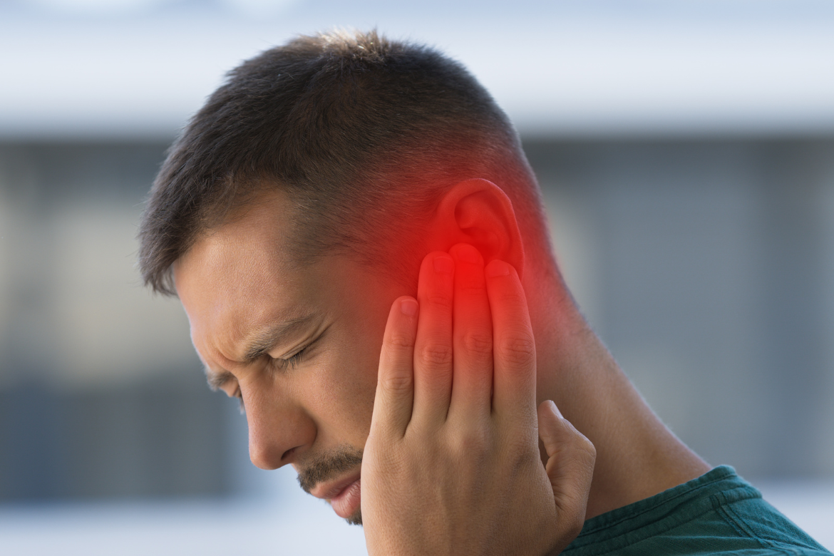 A man is looking down and wincing as though in pain. He's holding a hand to his left ear, which is highlighted in red, to suggest ear pain, infection or tinnitus.