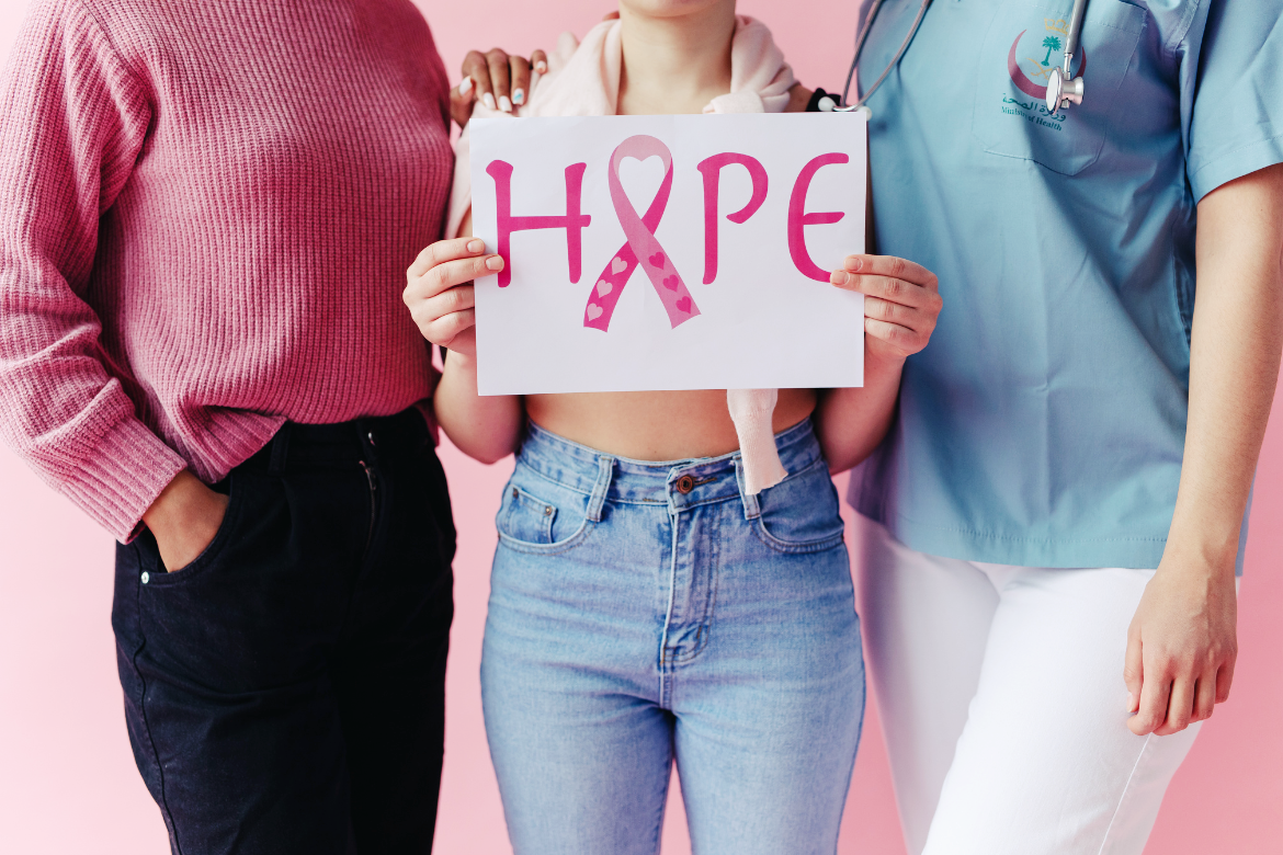 Three women, without showing their faces, are stood together. The middle one has a top around her shoulders and her front is bare. She's holding a sign in front of her reading "Hope" in pink lettering with a pink ribbon to signal breast cancer awareness and support.